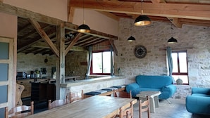 Een rustiek interieur met zichtbare houten balken, een stenen muur, een eethoek met houten meubels en een blauwe bank.
