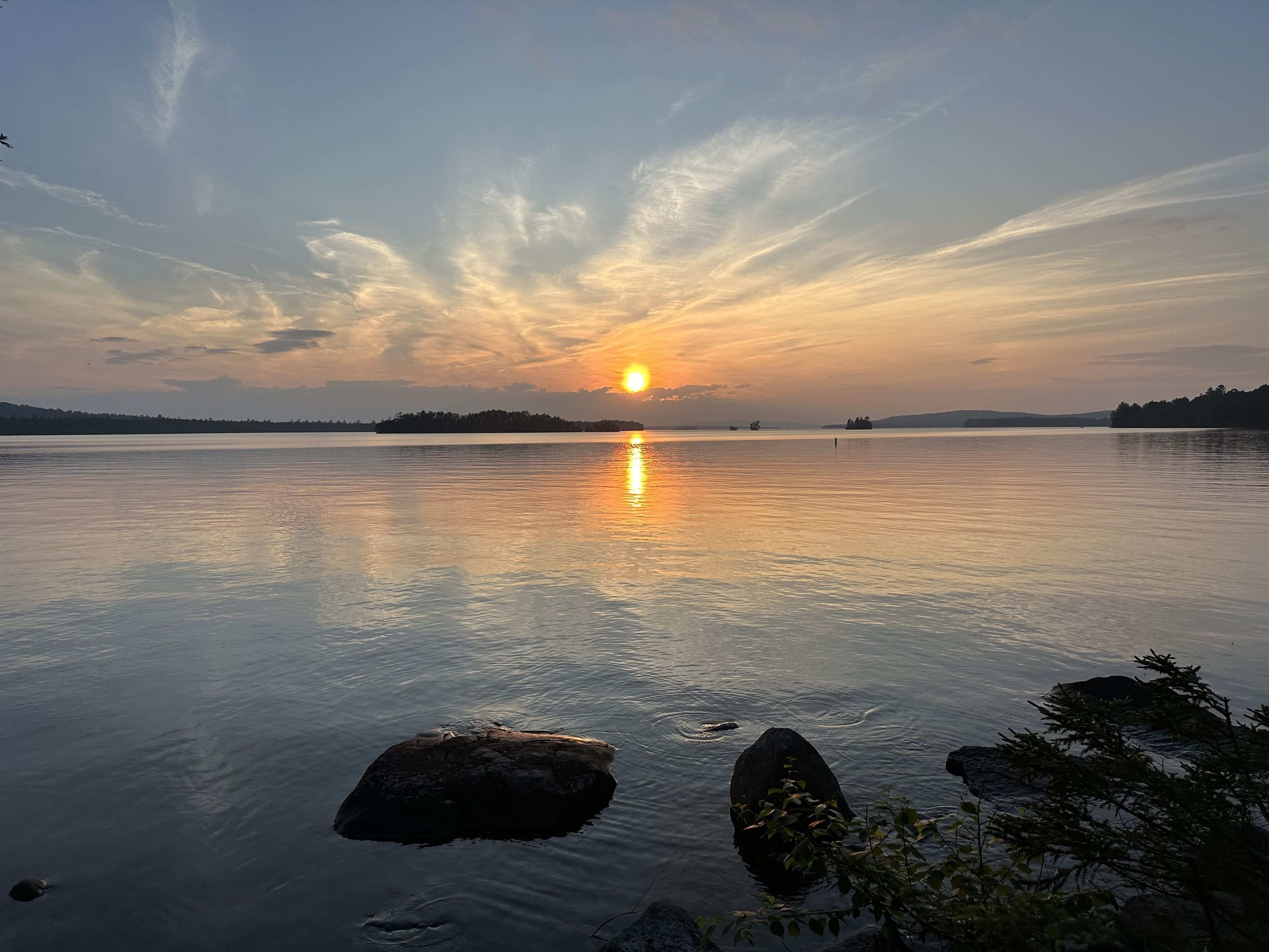Un llac tranquil a la posta de sol amb un cel clar, un sol visible i formes de terra a la distància.