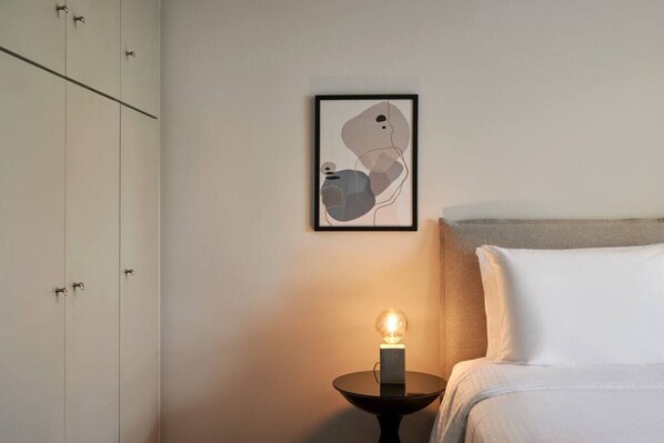 A bedroom with a bed, a bedside table with a lamp, a wardrobe, and a framed abstract artwork on the wall.