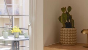 A cactus in a textured pot on a wooden shelf, a small table with a vase of yellow flowers, and a glass door leading to a balcony. - Urban Athéna Apartment,Pangrati (Athina)