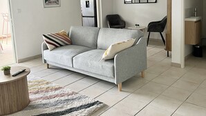 A modern living room with a grey sofa, a small round table, and a colorful rug. There is a door leading to another room, a chair, and a framed picture on the wall. - Appartement Duplex Bélize (Fort-de-France)