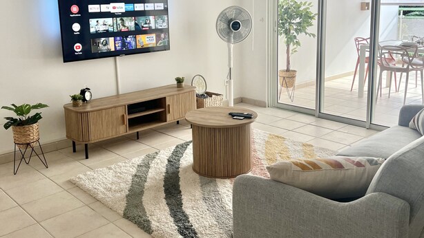 A modern living room with a flat-screen TV, wooden furniture, a fan, and a view through a glass door to an outdoor area. - Appartement Duplex Bélize (Fort-de-France)