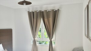 A bedroom with a bed, a chair, a window with curtains, and a hanging light fixture. - Appartement Duplex Bélize (Fort-de-France)