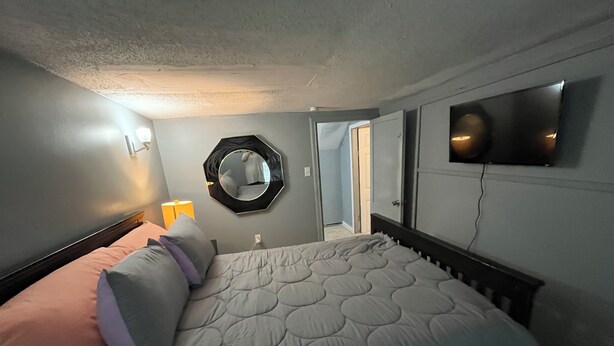 A bedroom with a bed, a wall-mounted TV, a mirror, and a door leading to another room.