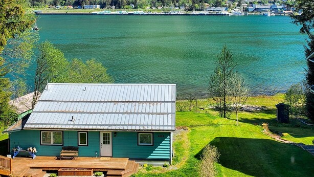 A lakeside cabin with a deck, surrounded by lush greenery and a mountain backdrop.