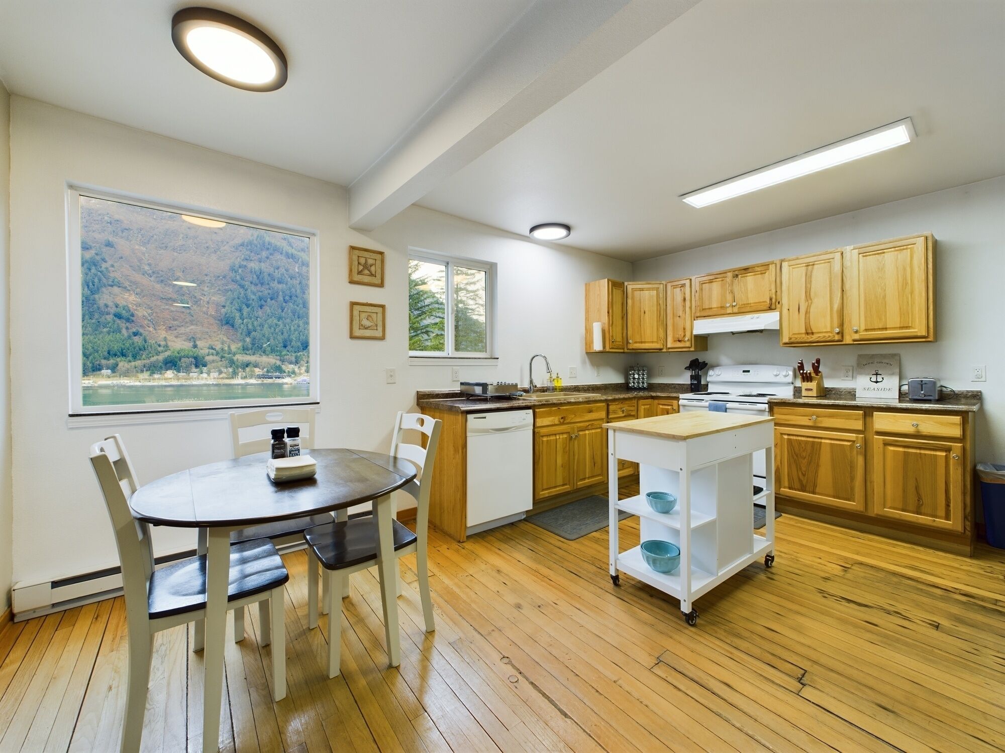 A modern kitchen with wooden cabinets, a dining table with chairs, and a view of a lake through large windows.