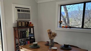 Uma área de refeições com mesa e cadeiras de madeira, uma estante e uma janela com vista para árvores.