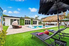 A modern house with a swimming pool, outdoor seating, and a thatched-roof structure. - • 1-Min Walk to Private Beach • Free Heated Pool • Tiki & Putting Green • Games (Pompano Beach)
