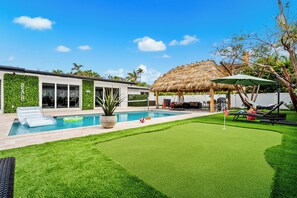 A modern house with a swimming pool, a thatched roof structure, and a well-maintained lawn. - • 1-Min Walk to Private Beach • Free Heated Pool • Tiki & Putting Green • Games (Pompano Beach)