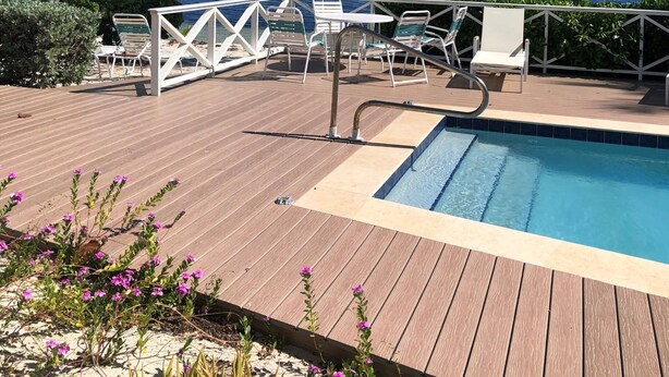 A wooden deck with a pool, white lounge chairs, and palm trees overlooking the ocean.