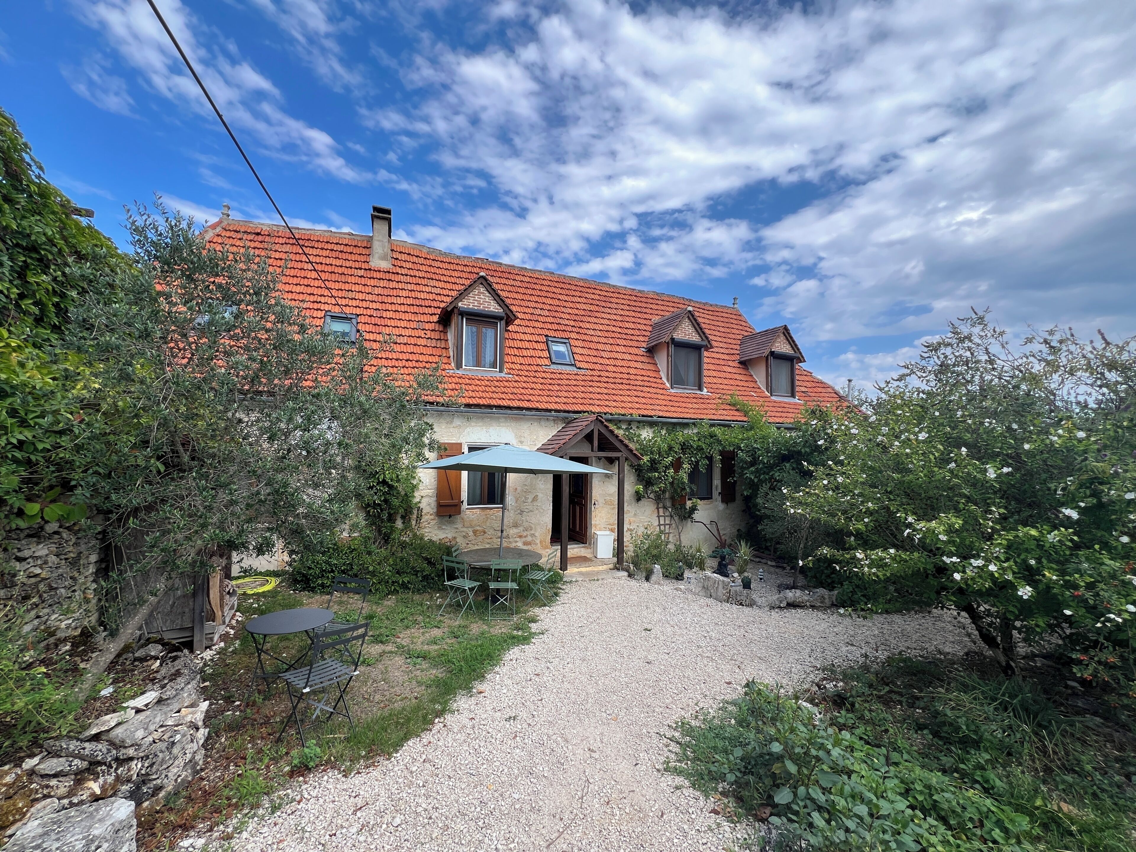 A house with a red-tiled roof, a gravel driveway, and a garden with various plants and a table with chairs.