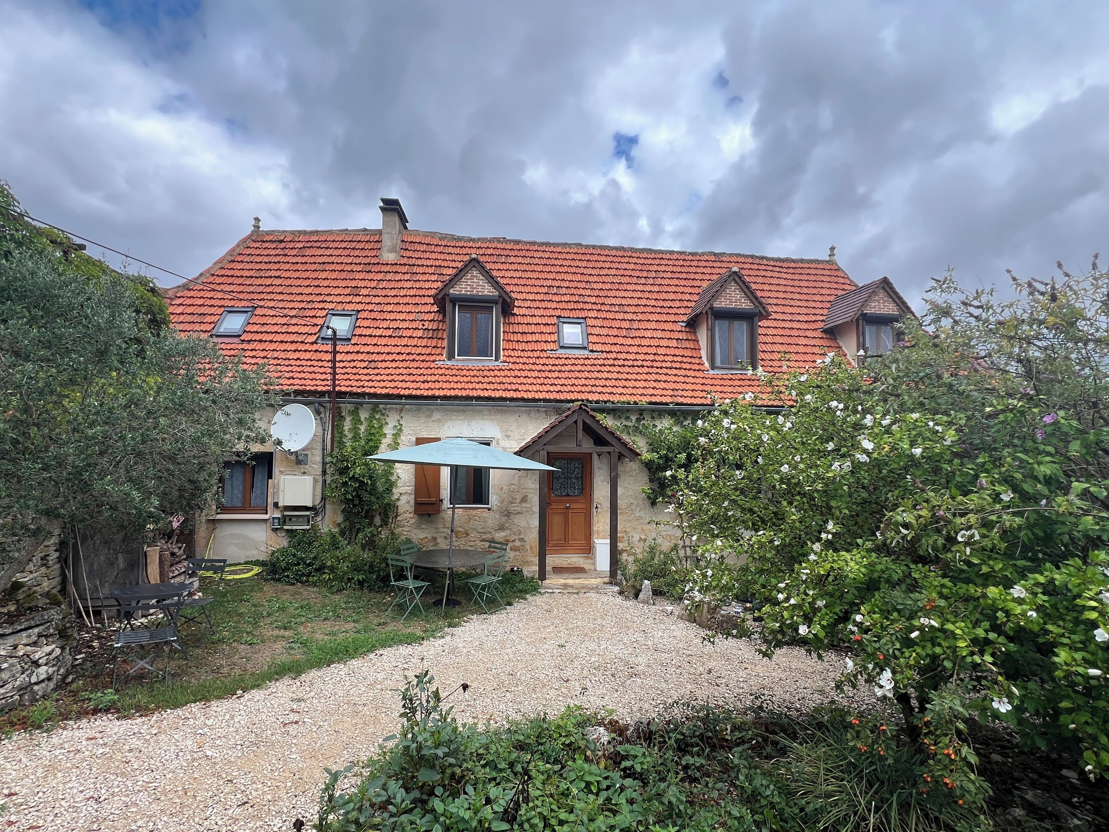 A house with a red-tiled roof, a gravel driveway, and a garden with various plants and flowers.