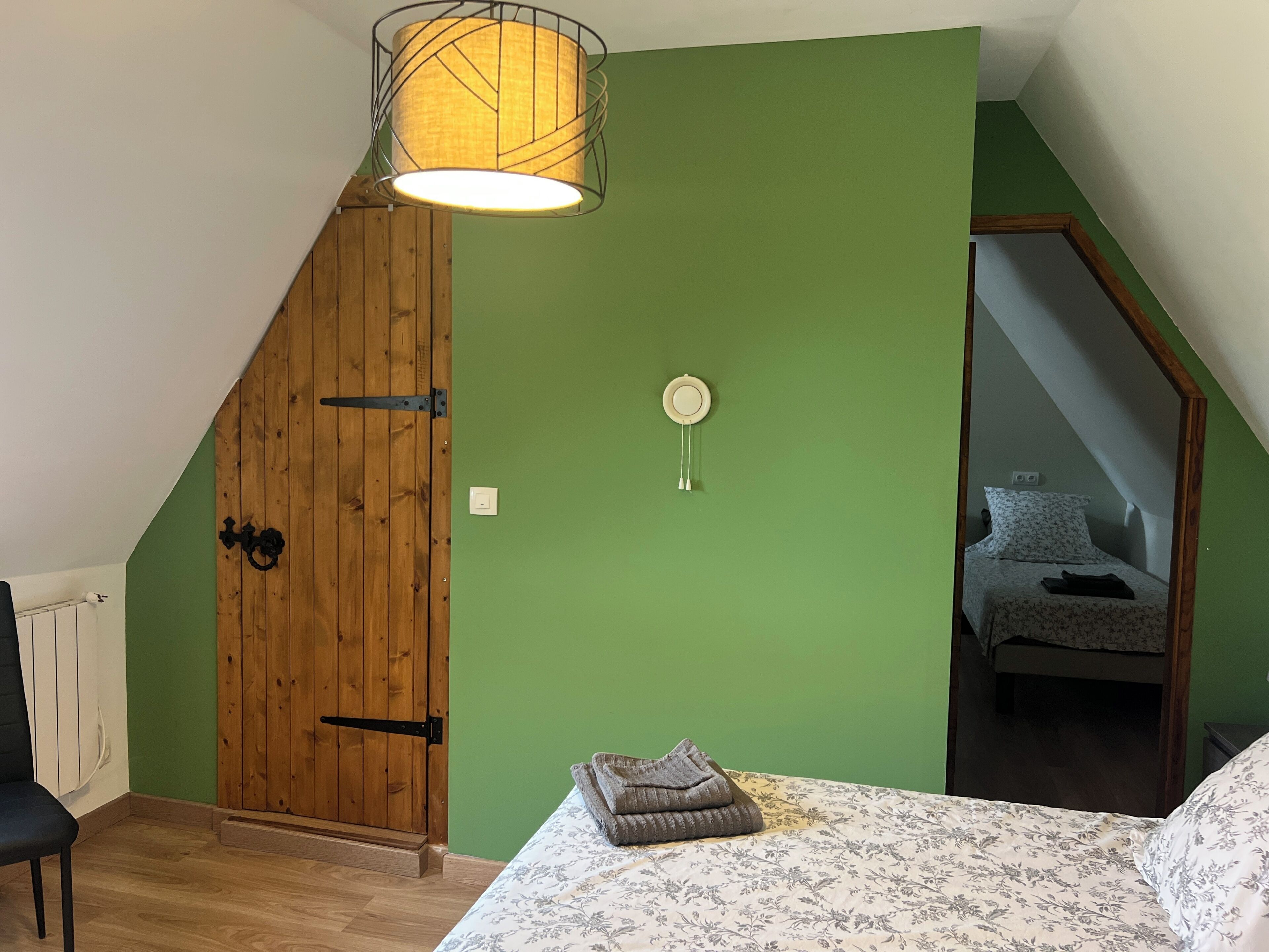 A bedroom with a sloped ceiling, a wooden door, a bed with a floral bedspread, a hanging light fixture, and a small bedside table.