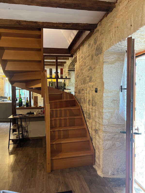 A wooden spiral staircase in a rustic interior with stone walls and a kitchen area visible in the background.