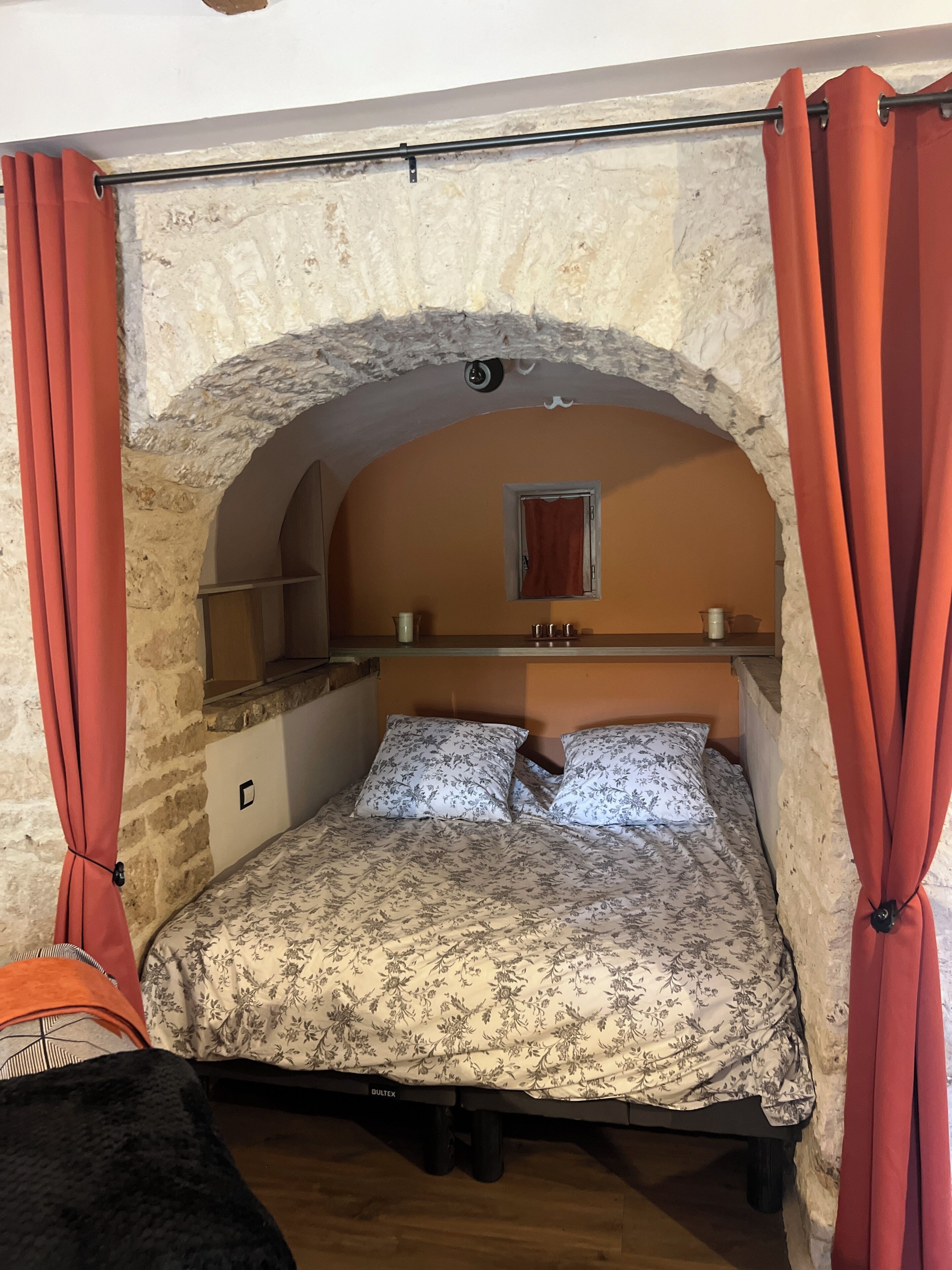 Un lit simple avec une couvre-lit à motifs floraux, un mur en pierre en arc de cercle, une petite fenêtre avec un rideau rouge et une étagère en bois au-dessus du lit.