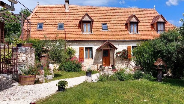 Une maison traditionnelle avec un toit recouvert de tuiles rouges, une façade en pierre et un petit jardin avec un chemin menant à l’entrée.