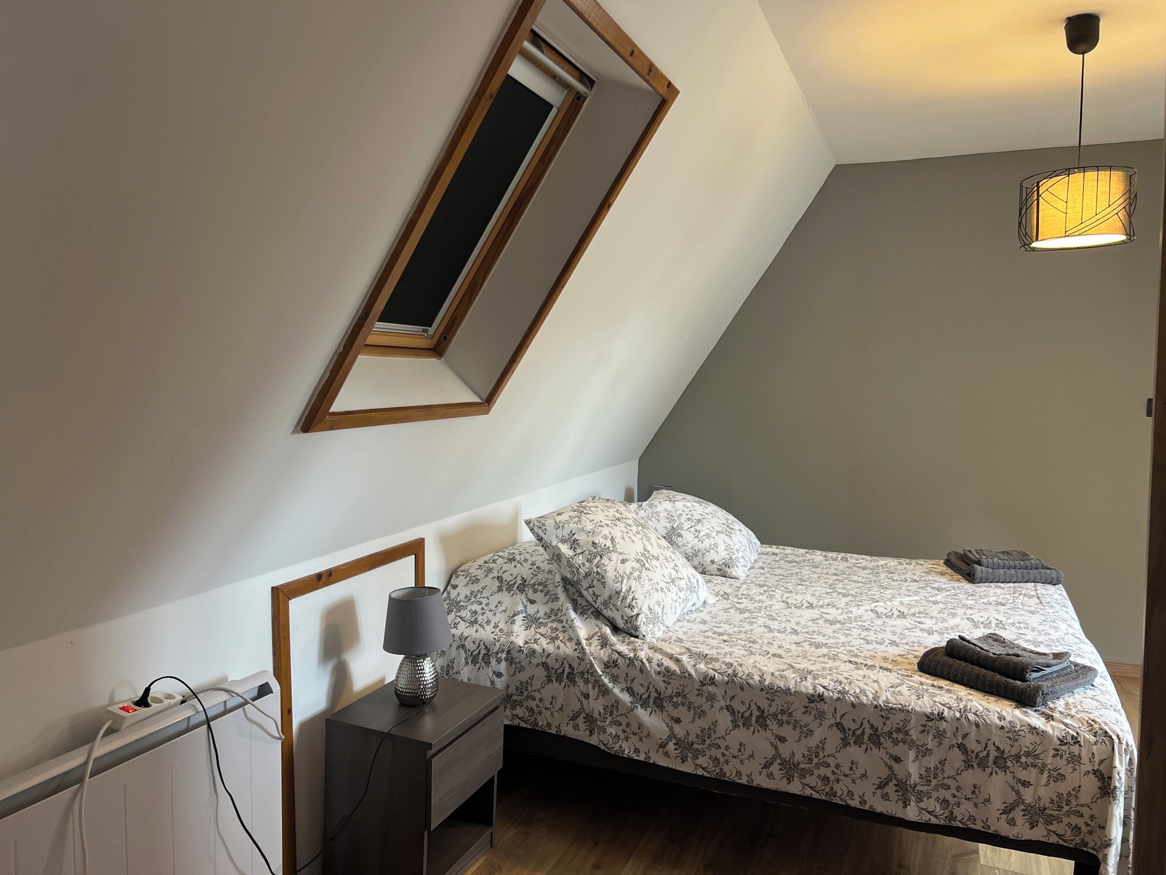 Une chambre avec un plafond en pente, un lit avec une literie à motifs floraux, une lampe de chevet, un miroir et un luminaire suspendu.