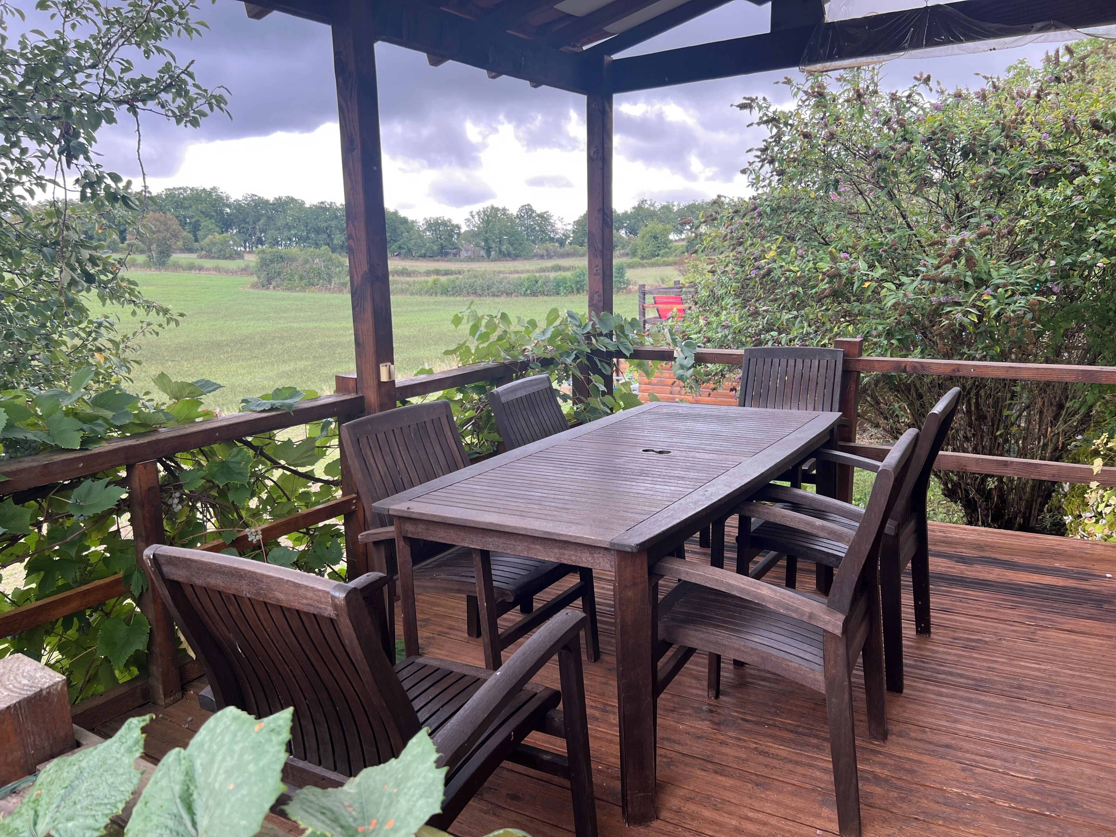 Een houten terras buiten, met een tafel en stoelen, omgeven door groen en uitzicht op een veld.