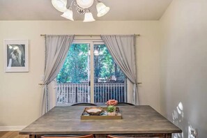 Un espace repas comprenant une table, des rideaux, un lustre et une fenêtre donnant sur des arbres, ainsi qu’un balcon.
