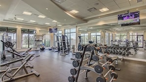 Un gimnasio espacioso con diversos equipos de ejercicio, ventanas amplias y una pantalla de televisión.