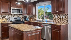 Uma cozinha com armários de madeira escura, bancadas de granito, eletrodomésticos de aço inoxidável e uma grande janela com vista para o verde.