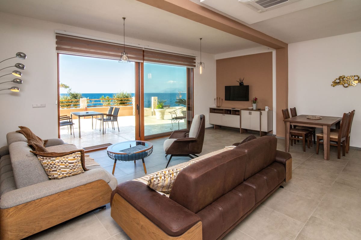 A modern living room with a sofa, armchair, and dining table. A large sliding glass door leads to an outdoor patio with a view of the sea.