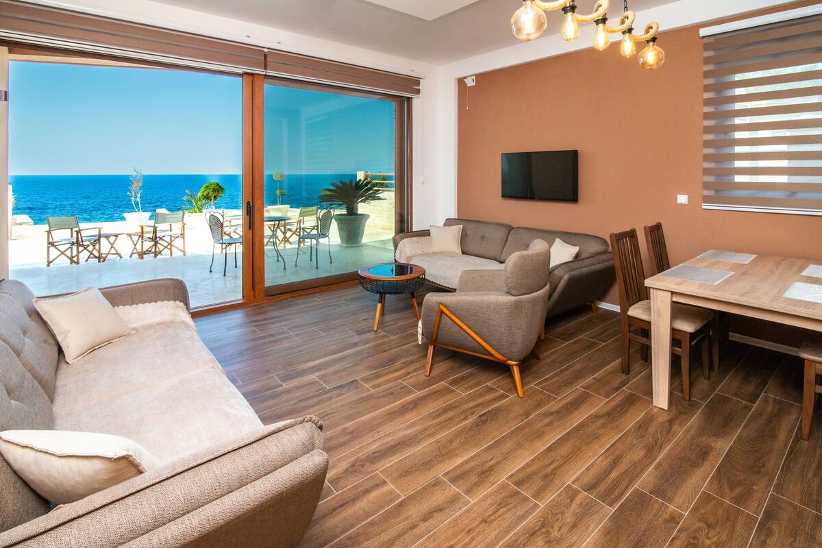 A spacious living area with a large sliding glass door leading to a balcony with a view of the sea. There is a sofa, a coffee table, and a dining table with chairs.