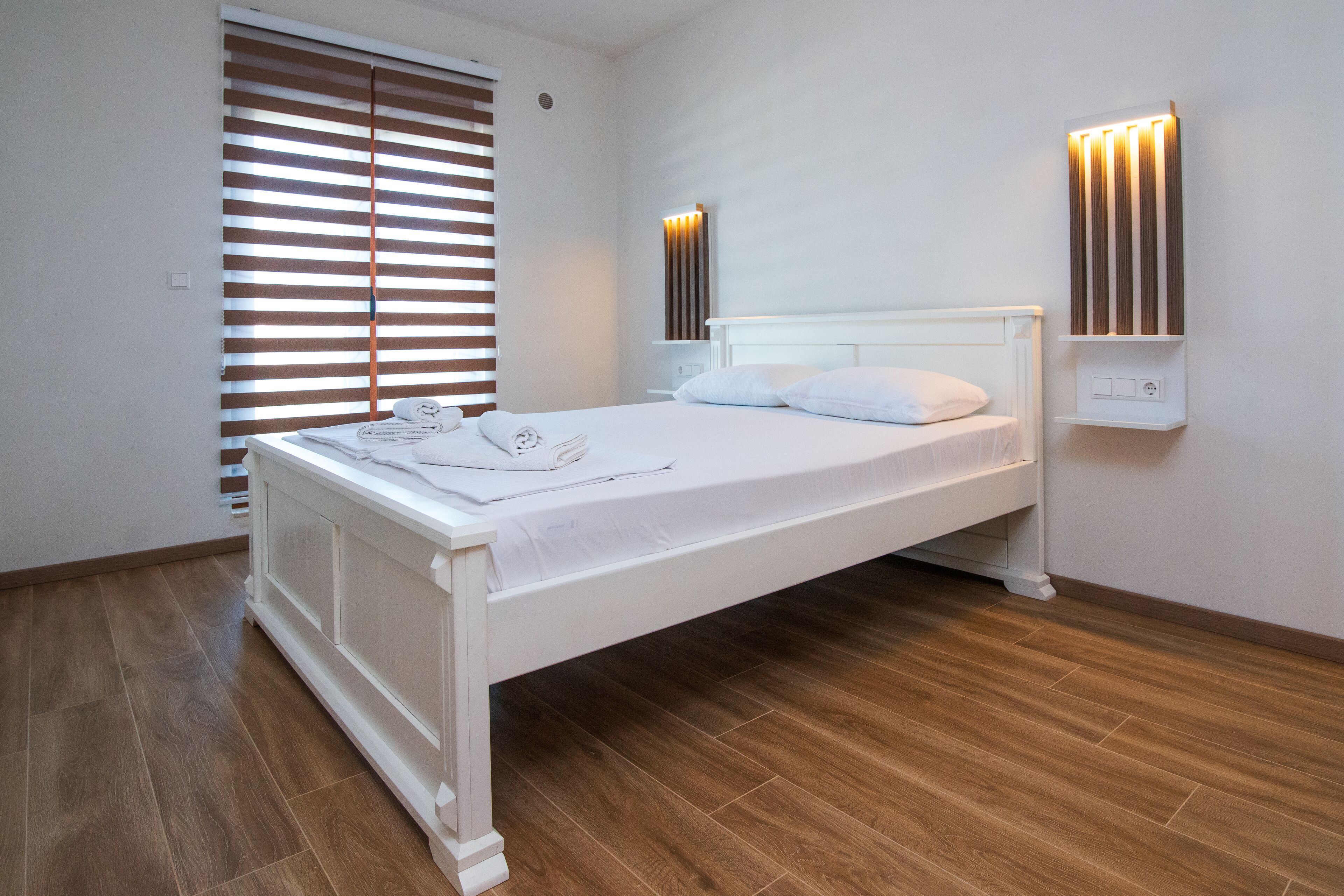 A neatly made bed with white linens, a wooden floor, and a wall-mounted light fixture.