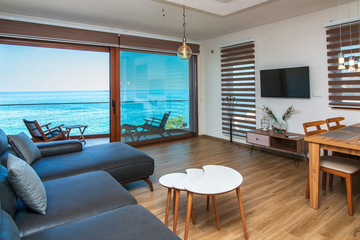 A modern living room with a large sliding glass door leading to a balcony overlooking the sea.