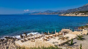 Una località balneare con lettini e ombrelloni su un pontile in pietra, con vista sul mare e sulle montagne.