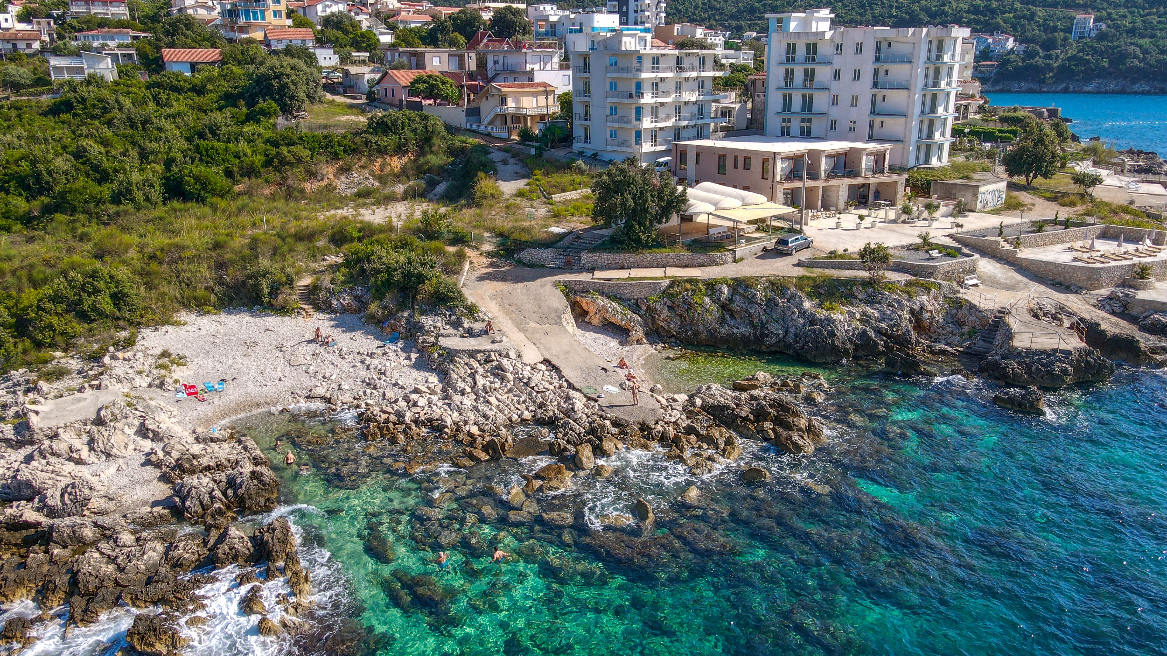Uma cidade costeira com praia de pedras, águas azul-claras e edifícios residenciais ao fundo.