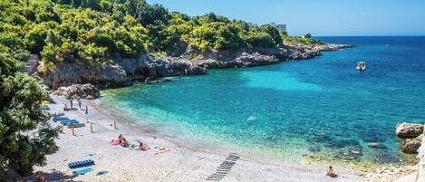 A beach with clear turquoise water, a rocky shoreline, and a building on a hill.