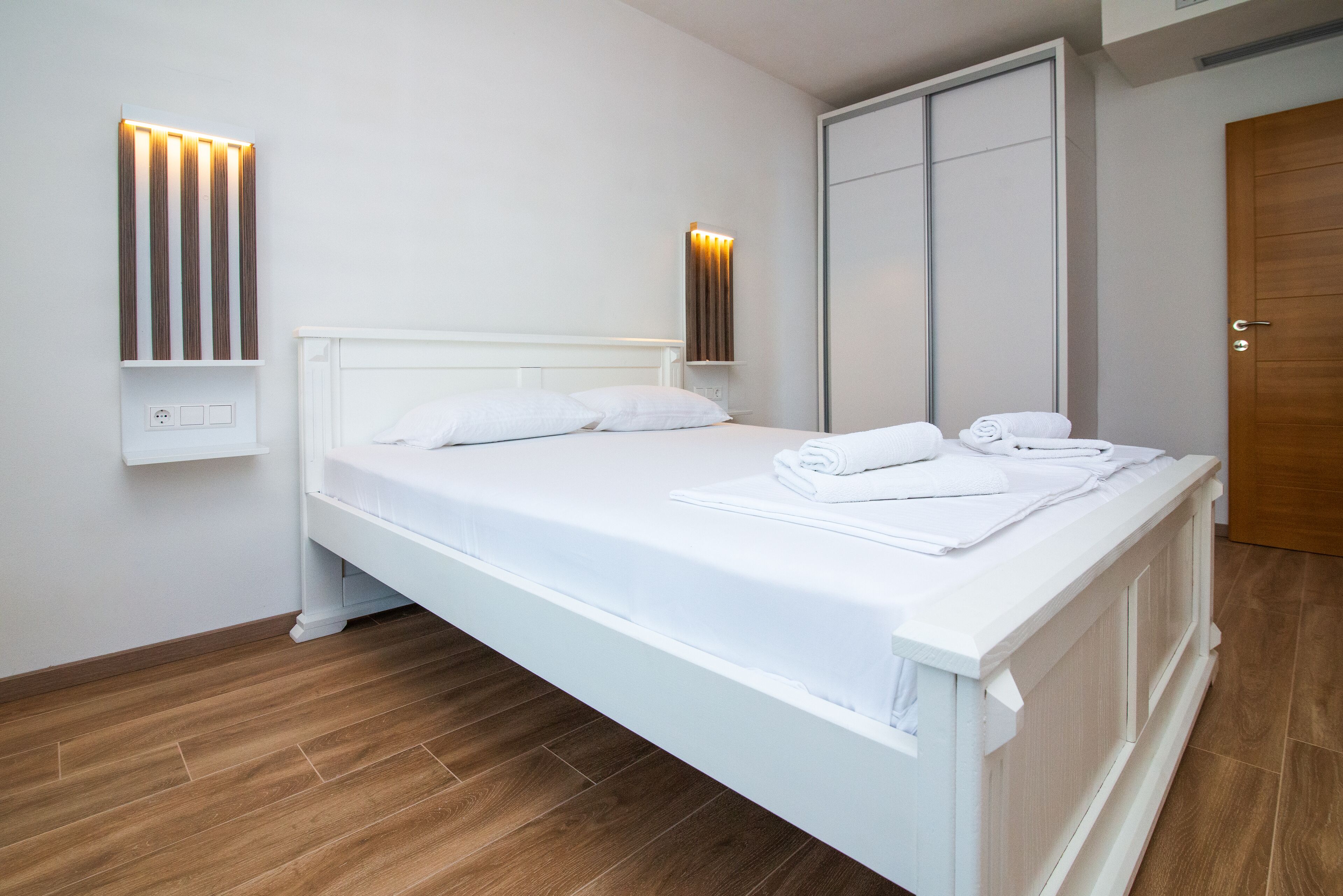 A neatly made bed with white linens and towels, a white headboard, and a wooden floor.