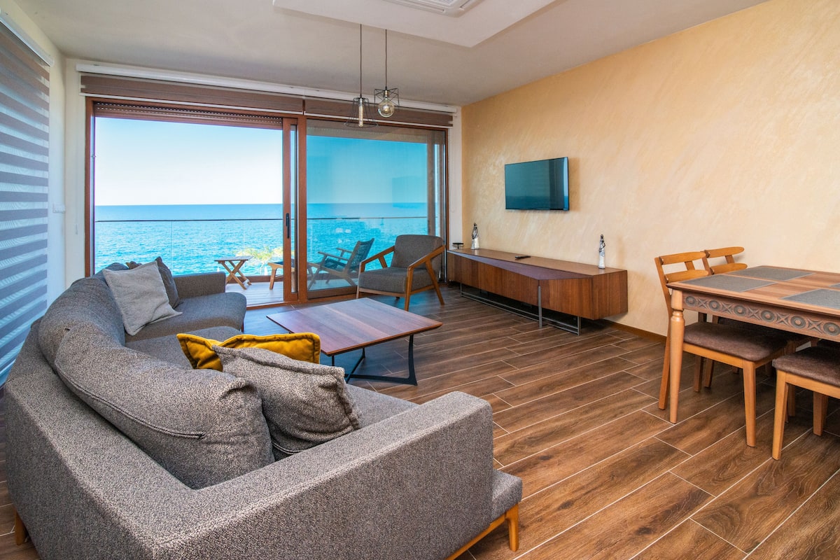 A modern living room with a large sofa, a coffee table, and a dining area with a wooden table and chairs. There is a TV mounted on the wall and a view of the ocean through large windows.
