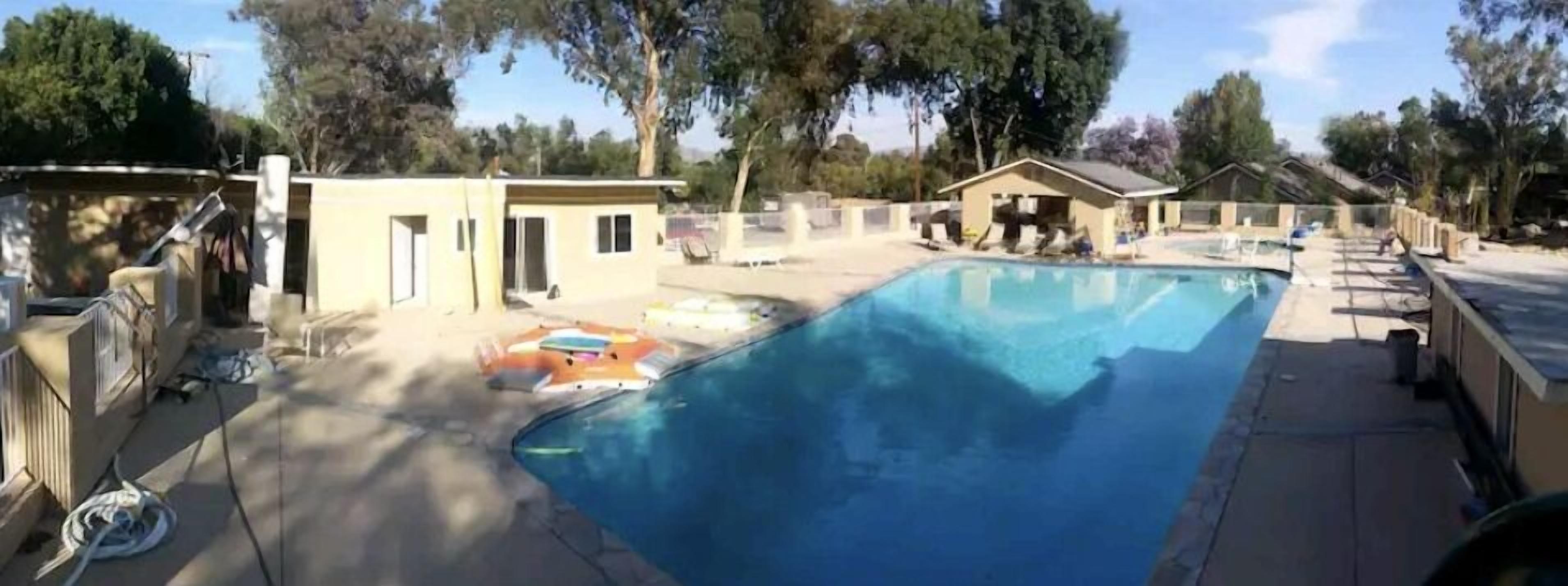 Una piscina con zona de solarium, rodeada de árboles y edificios pequeños.