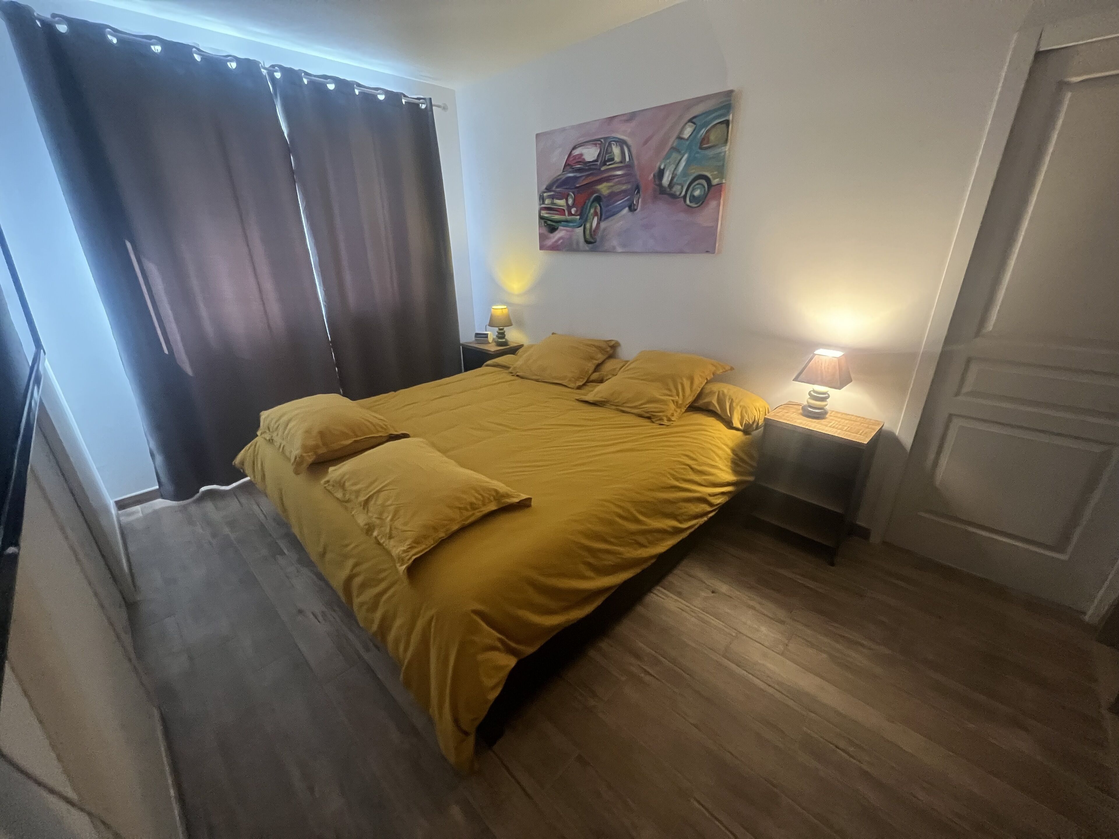 A bedroom with a large bed, two bedside lamps, a painting of cars on the wall, and a door on the right.