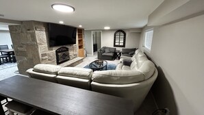 A living room with a stone fireplace, a flat-screen TV, a white leather sofa, a coffee table, and a dining area with a dark wooden table and chairs. - DEER CAVERN secluded 4 bedroom 2 bath house East Sandy base of Alta & Snowbird (Sandy)