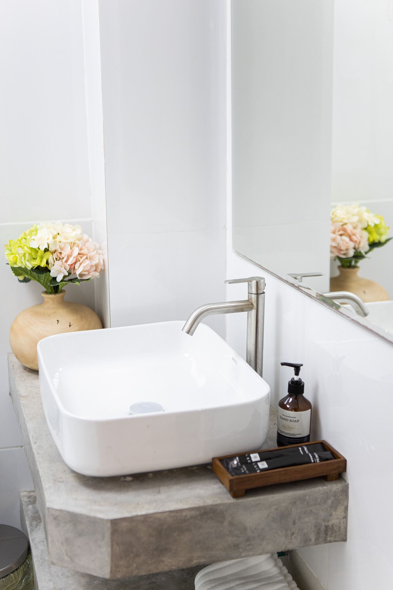 Ein Badezimmer mit einem weißen Waschbecken, einem Spiegel und einer Holze Vase mit Blumen.