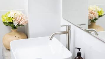 Ein Badezimmer mit einem weißen Waschbecken, einem Spiegel und einer Holze Vase mit Blumen.