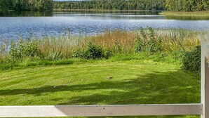 Un espace herbeux avec une clôture en bois au premier plan, une étendue d’eau et une forêt en arrière-plan.