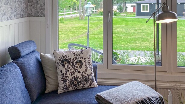 Une pièce avec un canapé bleu, un coussin à motifs et une vue sur une pelouse verdoyante et une maison, le tout visible à travers une grande fenêtre.