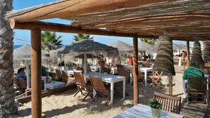 Un espace extérieur aménagé avec des tables et des chaises en bois, entouré de parasols en chaume et de palmiers.