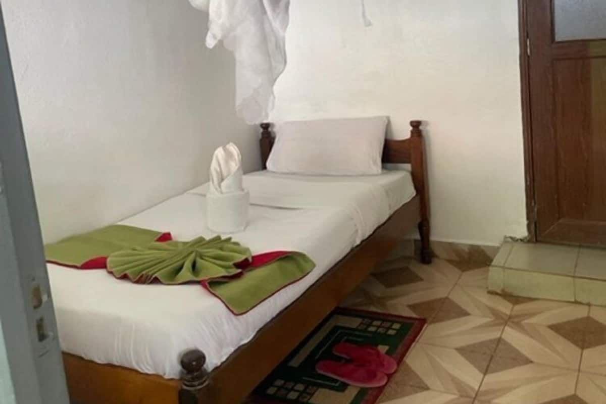 A single bed with a canopy, a wooden headboard, a bedside table with a white towel, and a red and green mat on the floor.