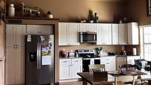 A spacious kitchen with white cabinets, a stainless steel refrigerator, a microwave, and a stove. There is a dining table with chairs and a wooden floor.