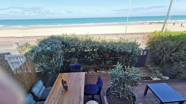 An outdoor seating area with a wooden table and chairs, surrounded by potted plants and a view of the beach.