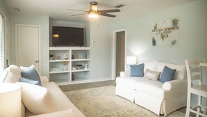 Ein Wohnzimmer mit einem weißen Sofa, einem weißen Sessel, einem Deckenventilator, einem Flachbildfernseher und einer wandmontierten Regal mit Dekorationen.