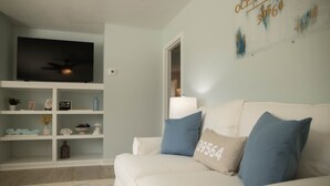 Ein Wohnzimmer mit weißem Sofa, blauen Kissen, einem Flachbildfernseher und einem Bücherregal.