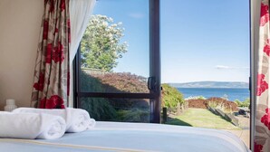 Un dormitorio con un ventanal grande, una cama con ropa blanca y vistas al mar y al follaje.