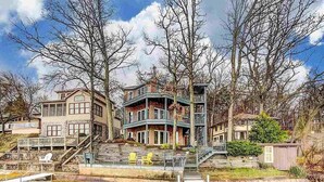 A multi-story house with a dock extending into a body of water, surrounded by trees. - Experience lakefront luxury with spectacular views on Lake James, Indiana! (Angola)