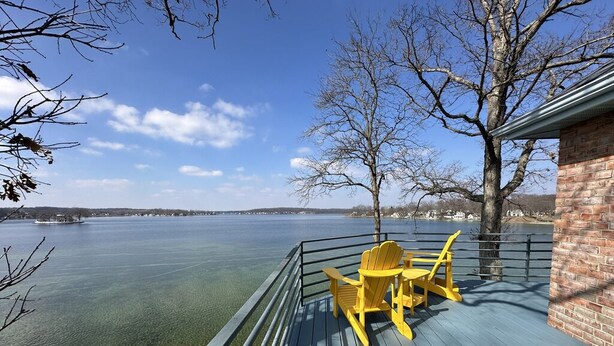 A deck with a table and chairs, overlooking a body of water and a distant shoreline. - Experience lakefront luxury with spectacular views on Lake James, Indiana! (Angola)
